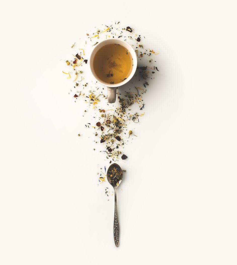 A cup of tea surrounded by scattered loose leaf tea and a spoon with tea leaves on a white background.